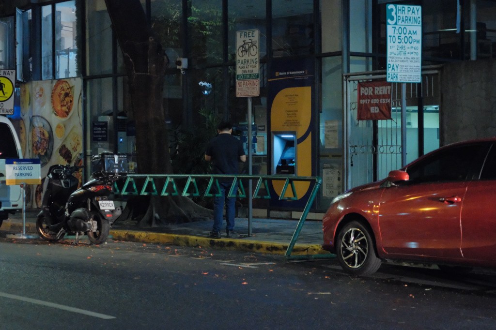 A bike rack in Salcedo Village, partially occupied, with a person using an ATM nearby. The setting includes vehicles parked along the street at night.