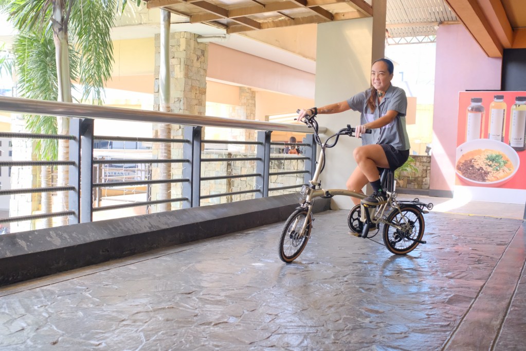 A person riding the Popcycle Friend e-bike on a smooth surface, with palm trees and a modern building interior in the background.