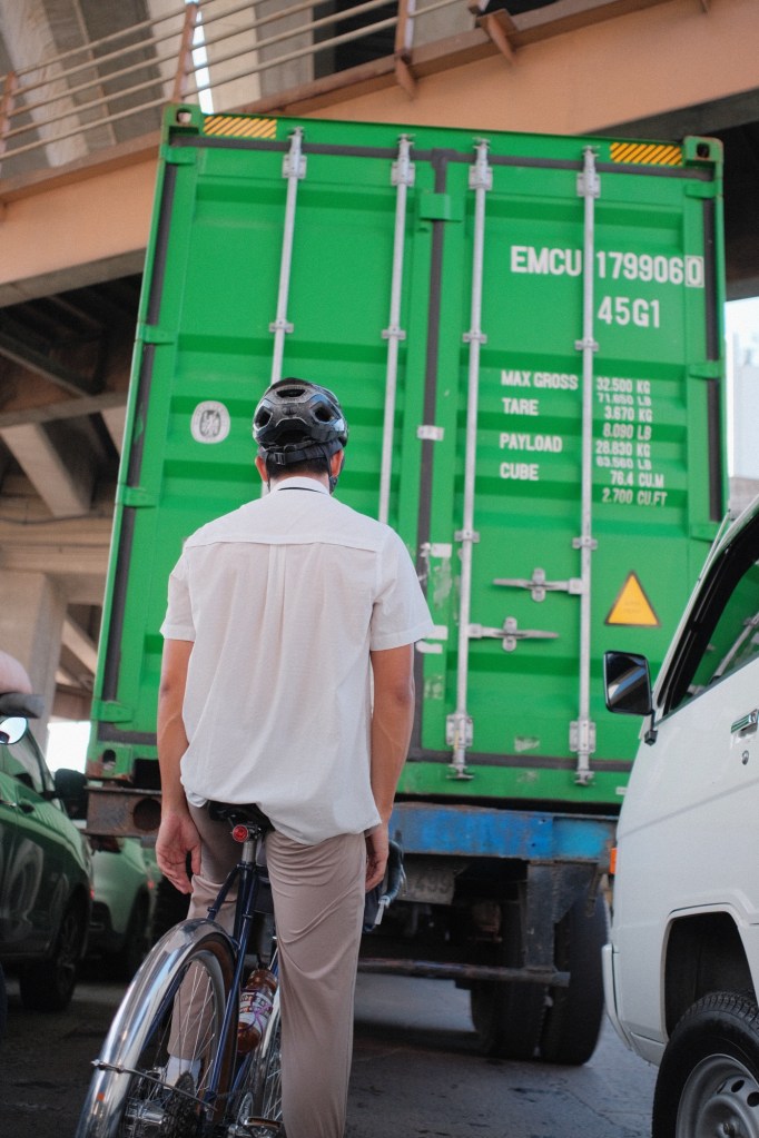 A cyclist in a white shirt and helmet going to Dampalit Megadike. He stands beside his bike, facing a large green shipping container truck in a congested urban area.