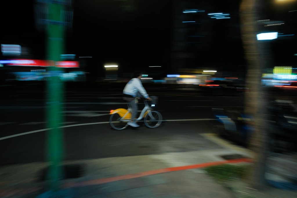 A blurred image of a cyclist riding a yellow bike at night, with city lights and street signs in the background. Example of mobility in Taipei