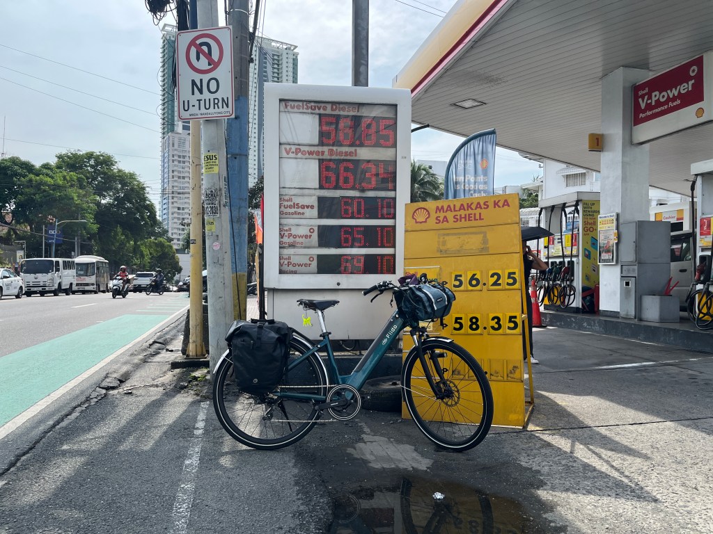 A bicycle parked next to a gas station displaying fuel prices, with a 'No U-Turn' sign nearby and a view of traffic on the road. bike commuting to san juan
