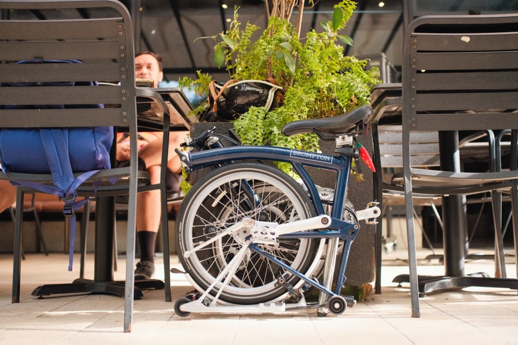 A folded Brompton C Line bike resting near a table with chairs, surrounded by plants in a cafe setting.