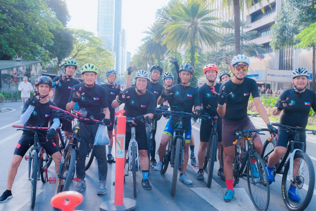 A group of cyclists from Deaf on the Road in matching black shirts with the Philippine flag, posing for a photo on a sunny street lined with palm trees and skyscrapers.