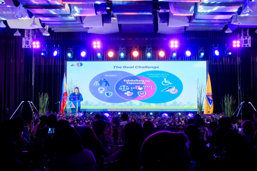 A speaker addresses an audience at an event titled 'The Dual Challenge,' highlighting issues related to women and persons with disabilities. A large screen displays relevant graphics and information, with colorful lighting in a formal setting. At the QC Women in Transport & Mobility Summit